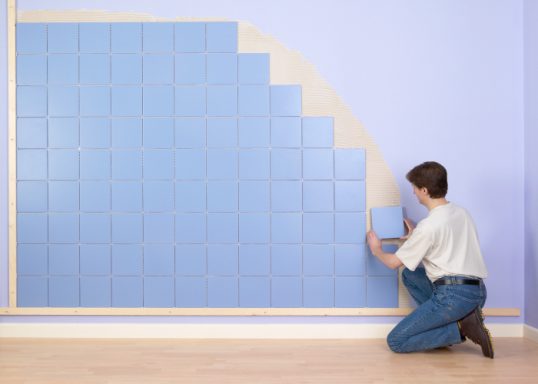 Ein Handwerker verlegt blaue Kacheln an einer Wand mit einer gewellten Holzleiste.
